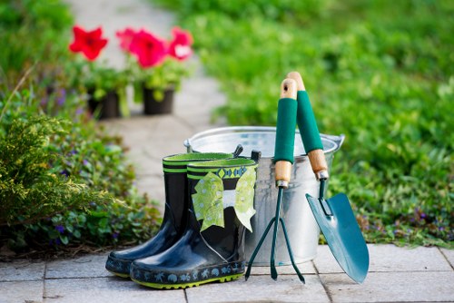 The image shows a garden scene with a paved pathway bordered by lush green grass and various plants. In the foreground, there are a pair of black rubber rain boots with a floral pattern, placed next to a white plastic bucket. Inside and beside the bucket, there are gardening tools, including a trowel with a teal handle, pruning shears, and a small hand rake. In the background, there are several vibrant pink and red flowers in pots, positioned on the garden pathway. The garden appears well-maintained with healthy foliage and a neatly arranged layout, suggesting an outdoor space suitable for gardening and lawn care services. The weather is clear, and the scene is lit by natural daylight, creating an inviting environment for outdoor gardening activities, aligning with professional gardening and landscaping services provided by Hedge Trimming Chelsea near the Chelsea area in London.