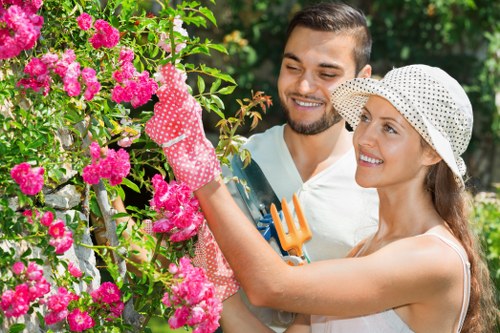 A man and a woman are working together in a well-maintained garden with lush greenery and vibrant pink flowering shrubs. The woman, wearing a white sunhat and gardening gloves, is pruning or trimming the flowering shrub using her hands, while the man, dressed in casual light clothing, stands beside her with a smile. The garden features a neatly edged flower bed, a lush green lawn in the foreground, and mature trees providing partial shade in the background. The overall scene suggests an outdoor gardening activity focused on plant maintenance and garden care, indicative of professional gardening services such as hedge trimming and shrub pruning offered by companies like Hedge Trimming Chelsea, rooted in an outdoor space that looks to be in or near Chelsea, London. Bright natural daylight highlights the colours and textures of the garden, creating a clean, inviting, and professional outdoor environment suitable for garden maintenance and landscaping services.
